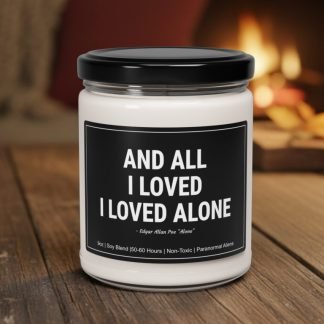 A white candle in a glass jar with a black lid on a wooden surface. The label quotes Edgar Allan Poe: "And all I loved, I loved alone." A cozy, lit fireplace is blurred in the background, conveying warmth and introspection.
