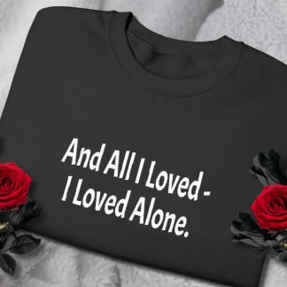A black sweatshirt with white text reading "And All I Loved - I Loved Alone" is laid on a soft gray blanket. Two red roses are placed beside it, conveying a somber tone.