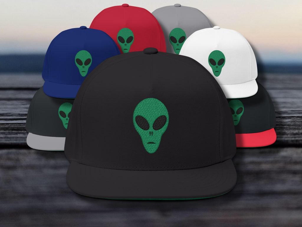 A collection of hats featuring a green alien face design. Prominently displayed in front is a black hat, surrounded by red, blue, gray, and white hats. The setting is a blurred outdoor scene.