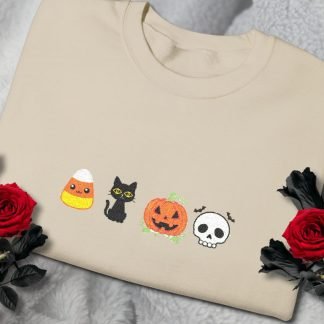 Beige sweatshirt with embroidered Halloween icons: candy corn, black cat, jack-o'-lantern, and skull. Red roses on a gray textured background. Spooky yet cozy.