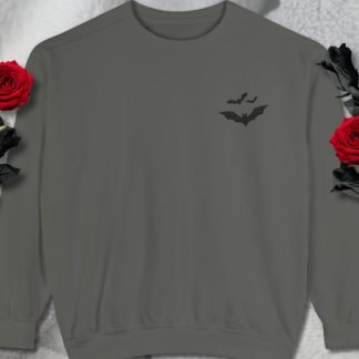 Gray sweatshirt with a small embroidered black bat design on the chest. Two red roses with black leaves frame the sweater against a soft, white textured background.