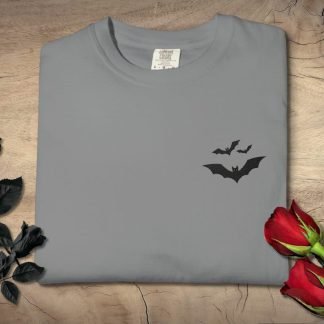 Folded gray T-shirt with embroidered black bat designs near the chest, surrounded by black roses on the left and red roses on the right, placed on wooden surface.