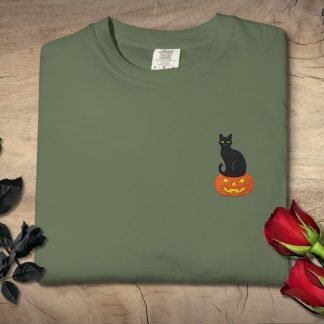 Green T-shirt with a small embroidered black cat sitting on an orange jack-o'-lantern. Black roses and red roses lie beside it on a wooden surface.