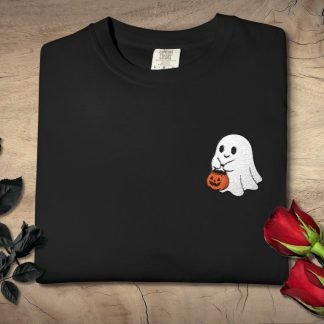 Black t-shirt with embroidered cute ghost holding a pumpkin design, placed on wood. Black roses to the left, red roses to the right, creating a spooky, romantic tone.