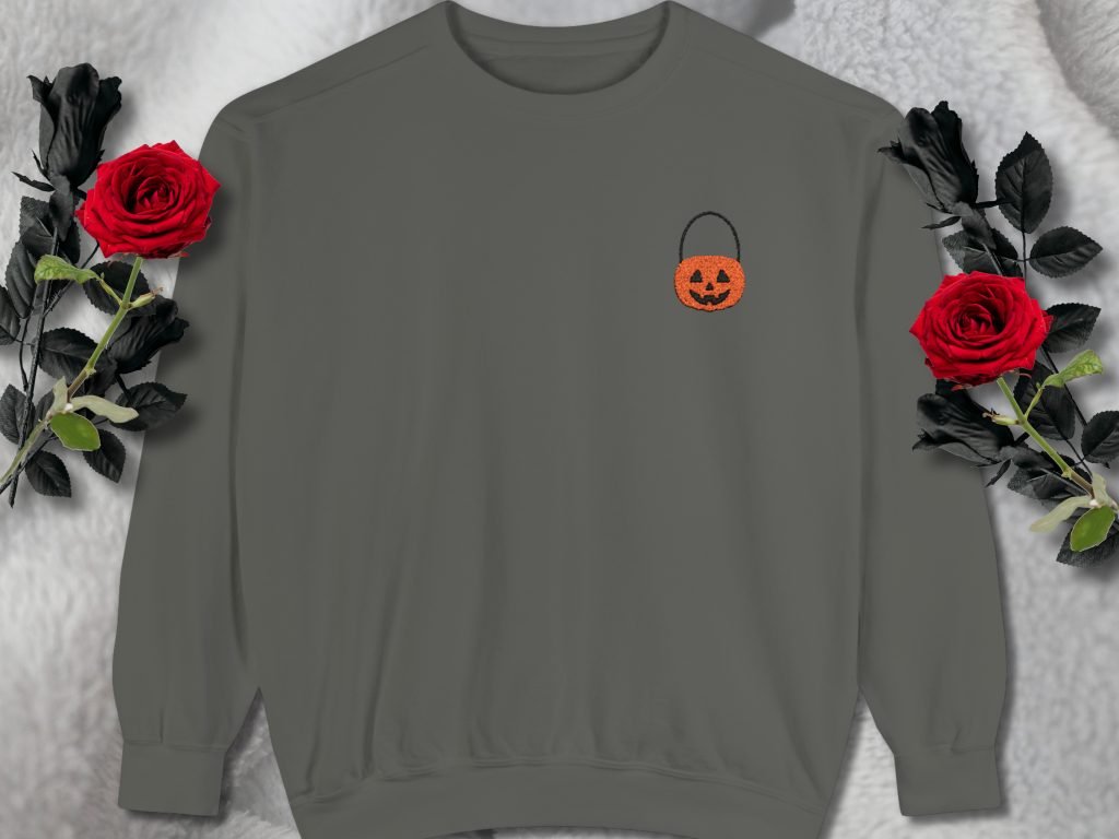 Gray sweatshirt with a small, orange Jack-o'-lantern embroidery on the left chest. Surrounded by two red roses on a soft, fluffy white background. Cozy and festive mood.
