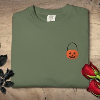 Folded olive green T-shirt with an embroidered orange pumpkin basket on a wooden background. Black roses on the left, red roses on the right.