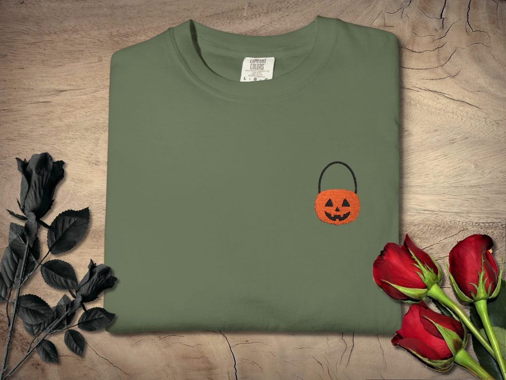 Folded olive green T-shirt with an embroidered orange pumpkin basket on a wooden background. Black roses on the left, red roses on the right.