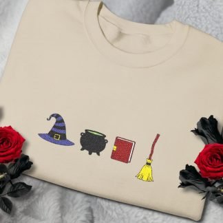 A beige sweatshirt with embroidered symbols: a witch hat, cauldron, book, and broom. Red roses on either side, set on a soft grey background. Cozy and mystical.