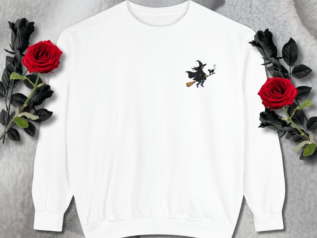 White sweatshirt featuring a small witch on a broomstick design on the chest. Surrounded by black roses with vibrant red blooms on a soft, gray background.