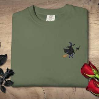 A folded green T-shirt featuring a small embroidered witch on a broomstick is placed on a wooden surface. Beside it are black and red roses.