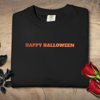 A black embroidered "Happy Halloween" T-shirt with orange text is neatly folded on a wooden surface. Beside it are black and red roses, adding an elegant touch.