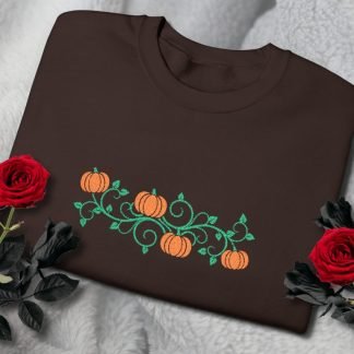 Brown sweatshirt with embroidered orange pumpkins and green vines, placed on a soft gray blanket. Surrounded by vibrant red roses, creating a cozy, autumnal feel.
