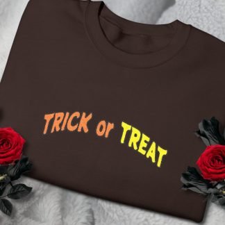 Folded brown sweatshirt with "TRICK or TREAT" in orange and yellow text, surrounded by black roses with vibrant red blooms, on a light gray textured fabric.