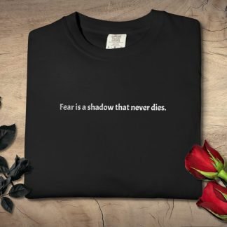 Folded black T-shirt with white text saying, "Fear is a shadow that never dies." Placed on a wooden surface, surrounded by black and red roses. Mood is contemplative.