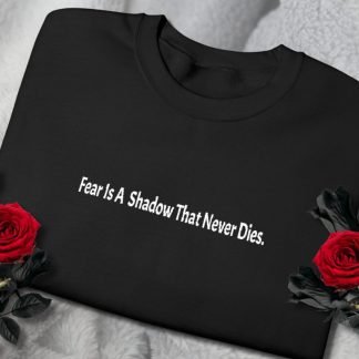 Folded black sweatshirt with white text, "Fear Is A Shadow That Never Dies," placed on gray fabric. Two red roses with dark leaves flank the shirt, creating a moody tone.