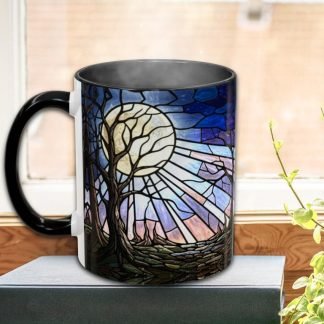 A black mug features a stained glass design of a tree with a large moon and radiant sky in blues and purples. It sits on a book by a sunlit window.