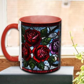 A mug with a stained glass design featuring red and purple roses sits on a book by a sunny window. The scene conveys warmth and vibrancy.