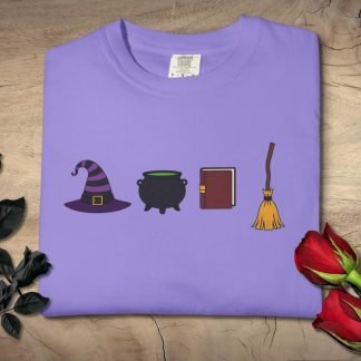Folded lavender t-shirt embroidered with a witch-themed design: hat, cauldron, book, broom. Surrounded by black roses and red roses, on a wooden surface.