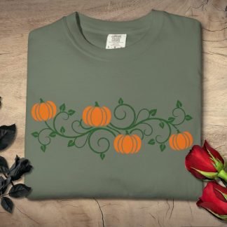 Gray T-shirt embroidered with orange pumpkins and green vines. Laid on a wooden surface, flanked by black roses on the left and red roses on the right. Cozy and autumnal.