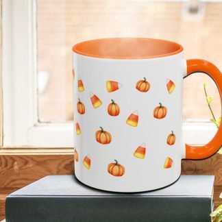 A white mug with an orange handle and interior features a pattern of small pumpkins and candy corn. It rests on a book by a sunlit window, creating a cozy autumn vibe.