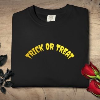 A black T-shirt embroidered with "Trick or Treat" in yellow, playful font. It rests on a wooden surface, flanked by black roses on the left and red roses on the right, evoking a spooky yet festive Halloween vibe.