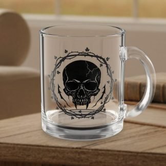 A clear glass coffee mug features a black skull design encircled by a thorny vine. It sits on a wooden table with books and a couch in the background, creating an edgy, cozy atmosphere.