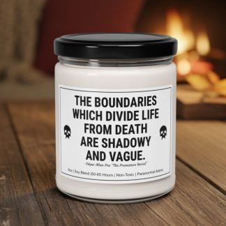 A white candle in a glass jar with a black lid sits on a wooden table. The label reads, "The boundaries which divide life from death are shadowy and vague" with a skull icon. A warm, blurred fireplace is in the background.