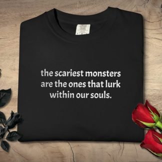A black shirt with the quote "the scariest monsters are the ones that lurk within our souls" in white text. Red and black roses frame the shirt on a wooden background, conveying a reflective and somber tone.
