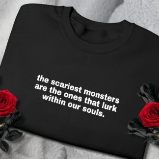 Black sweatshirt with white text: "the scariest monsters are the ones that lurk within our souls." Laid on gray fabric, flanked by two red roses.
