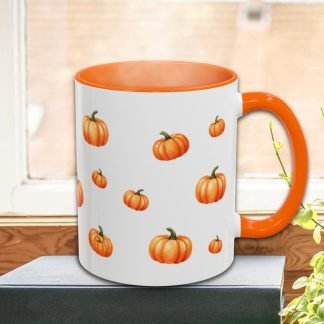 A white mug with an orange handle and rim is decorated with small, scattered pumpkin illustrations. It sits on a book by a window, next to green plants.